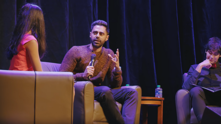 Hasan Minhaj sitting in an armchair on a stage, in conversation with two student interviewers.