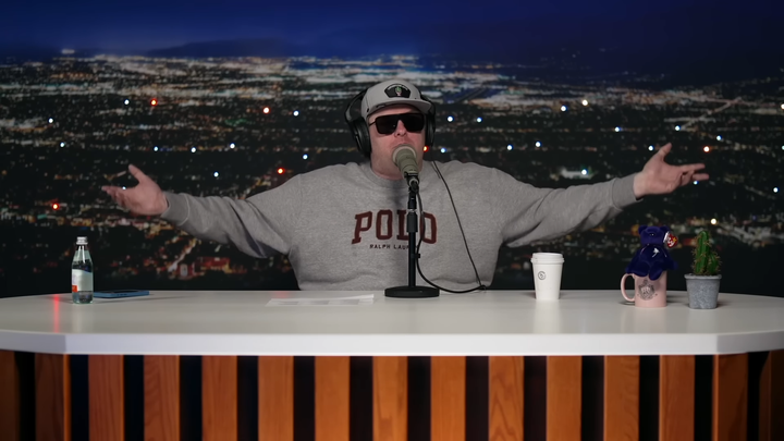 Tim Dillon sitting at his desk in his studio, wearing sunglasses, holding his arms out. 